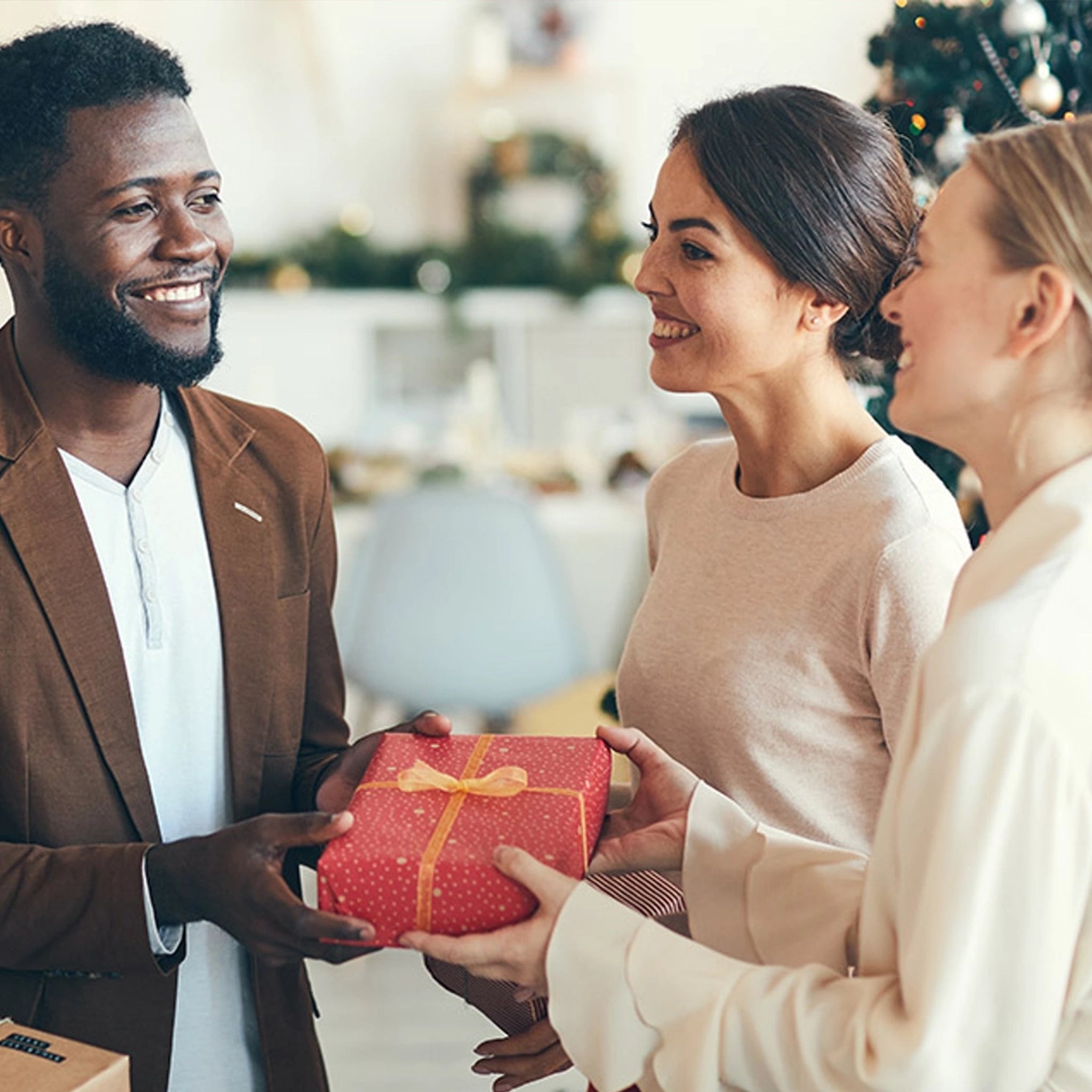 zwei lächelnde und creme-weiss gekleidete Frauen überreichen einem erfreuten Mann mit braunen Sakko und weissem Shirt ein in rotem Papier verpacktes Geschenk.