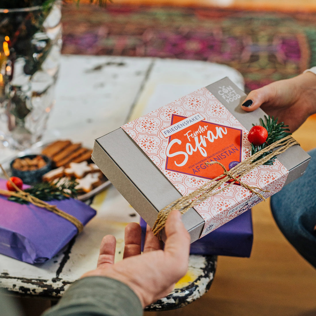 Eine weibliche Hand überreicht einer männlichen Hand ein festlich dekoriertes Conflictfood Paket Safran aus Afghanistan. Im Hintergrund auf einem Tisch sind Weihnachtsplätzchen und blaue verpackte Geschenke