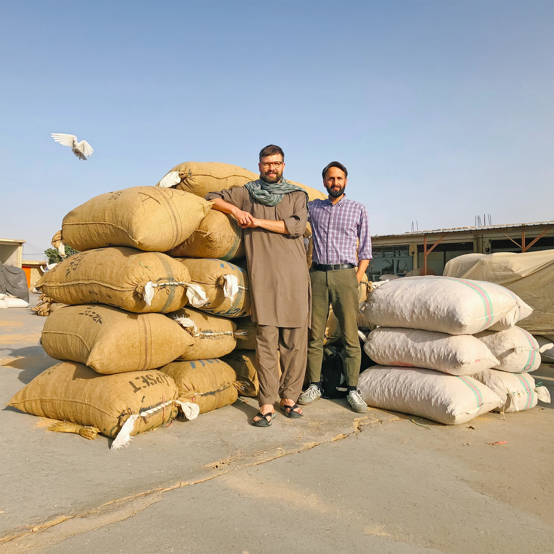 Gernot Würtenberger und Salem El-Mogaddedi von Conflictfood stehen vor einem Turn mit Säcken am Markt in Afghanistan, dahinter fliegt eine Friedenstaube