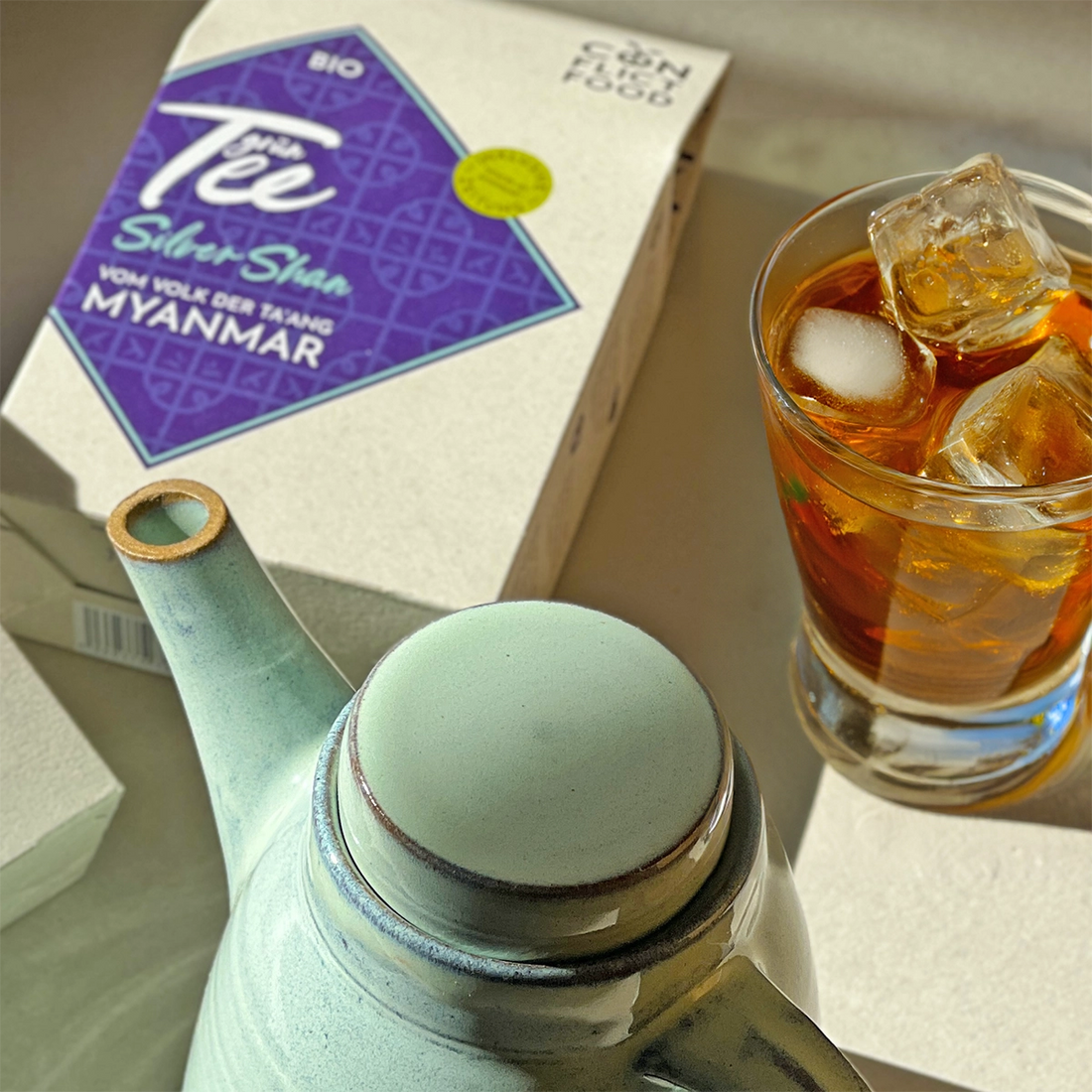 Eine Packung Conflictfood Silver Shan Grüntee neben einer Teekanne und einem Glas voll Eistee