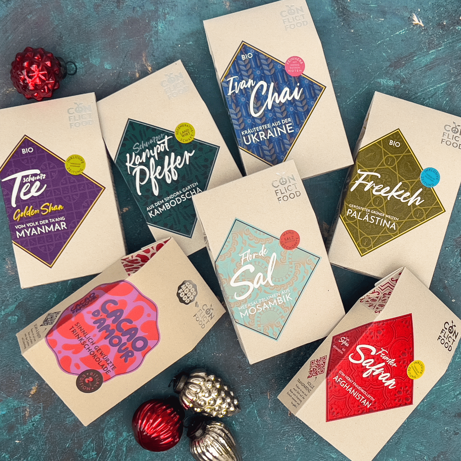 Sieben Conflictfood Päckchen Ivan Chai, Schwarztee Golden Shan, Feinster Safran, Schwarzer Kampot Pfeffer, Freekeh, Flor de Sal, Cacao DAmour liegen auf grünem Hintergrund und dazu weihnachtliche Dekoration in rot und silber