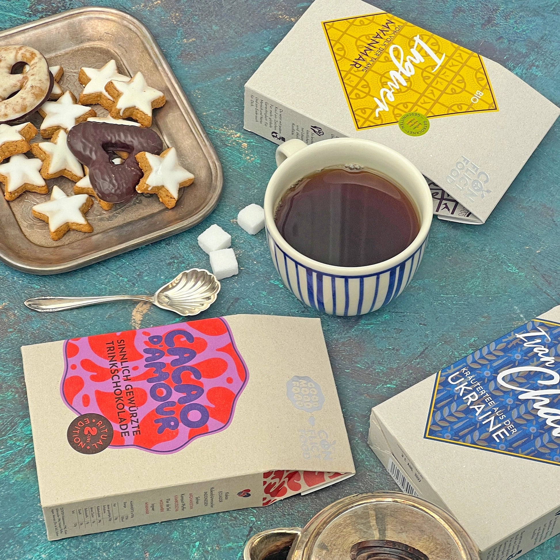 Drei Conflictfood Päckchen Ivan Chai aus der Ukraine, Ingwer aus Myanmar und Cacao D`Amour Trinkschokolade liegen auf blauen Grund. Zusammen mit Tasse Tee, Zuckerwürfel, und Weihnachtsplätzchen auf Silbertablett und Silberlöffel