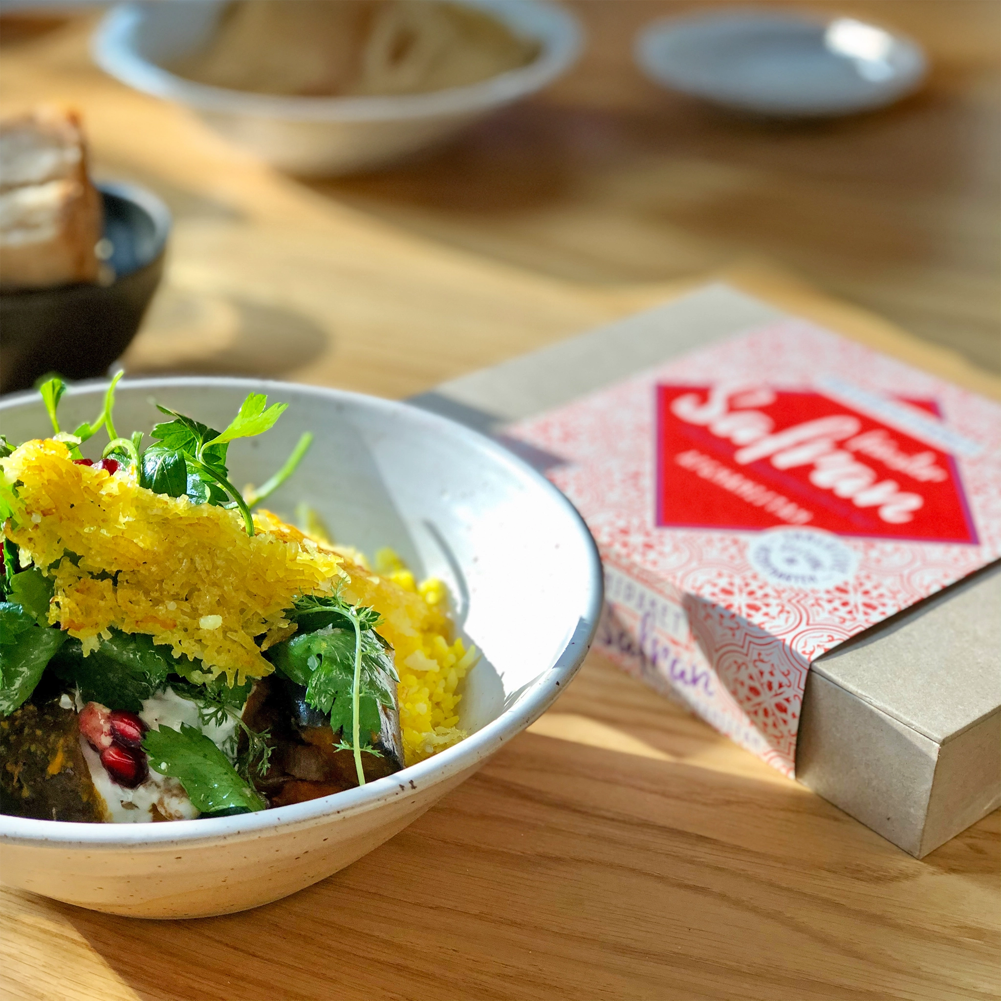 Conflictfood Paket Safran aus Afghanistan auf Holztisch. Daneben weisse Schüssel mit Safranreis und Salat. Im Hintergrund blaue Schale mit Brot