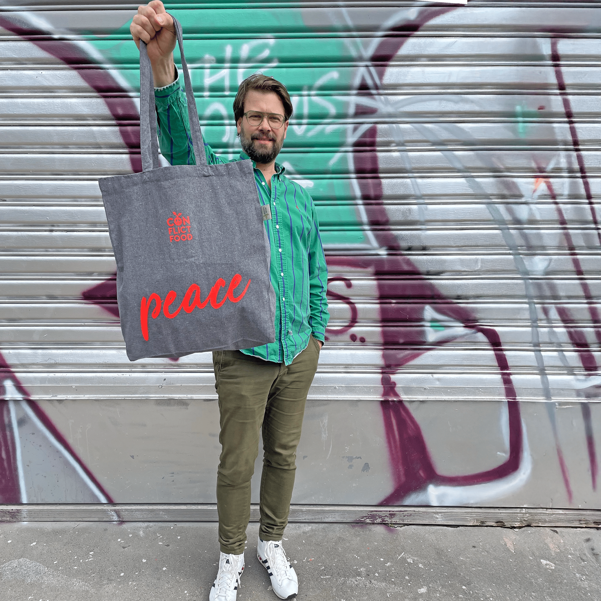 Ein Mann mit Brille und grüner Hose und grünem Hemd und weissen Sneakers steht vor einem Graffiti besprühtem Rolltor. Er hält einen grauen Baumwollbeutel von Conflictfood in der Hand. Mit rotem Conflictfood-Logo und der Aufschrift PEACE