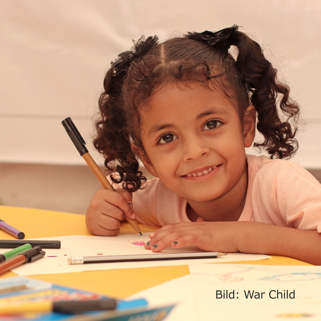 Ein junges Mädchen malt mit bunten Stiften auf einem Blatt Papier und lächelt dabei in die Kamera. Bild: War Child