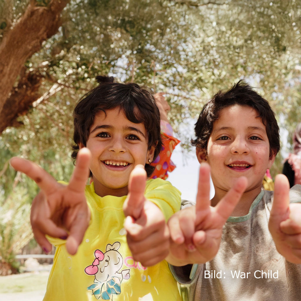 Zwei junge Buben formen mit ihren Fingern ein Peace Zeichen und schauen in die Kamera. Bild: War Child