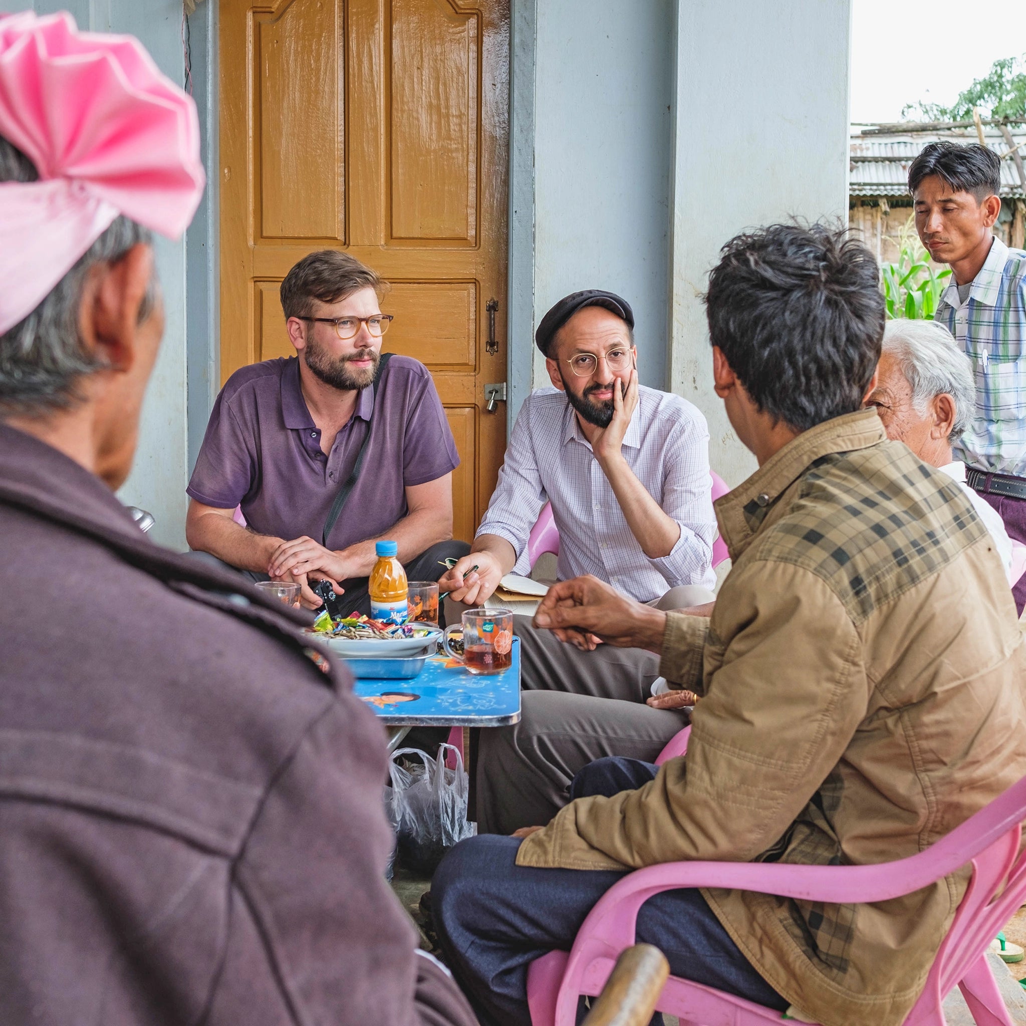Die Gründer von Conflictfood am blauen Tisch sitzend auf rosa farbigen Stühlen inmitten eines Gespräches mit mehreren männlichen Teeproduzenten in Myanmar