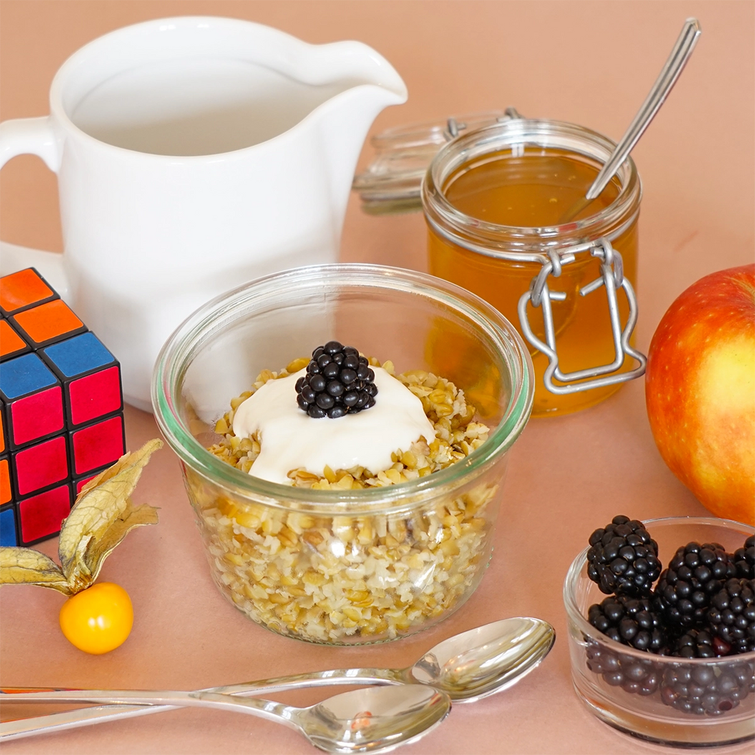 Ein Glas gefüllt mit Freekeh und Joghurt und daneben Beeren, ein Apfel und ein Milchkännchen sowie Löffel und ein Rubix-Würfel