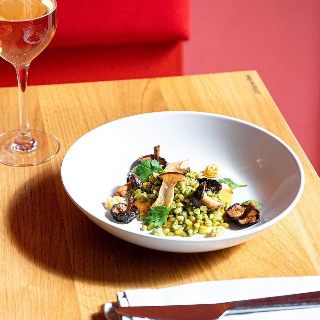 Auf einem Tisch steht ein Teller mit Freekeh Risotto und Pilzen, daneben ein Glas Wein