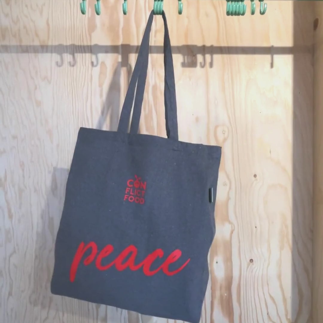 Video laden: Grauer Baumwollbeutel mit rotem Conflictfood-Logo und der Aufschrift PEACE hängt an einem grünen Garderobenhaken 