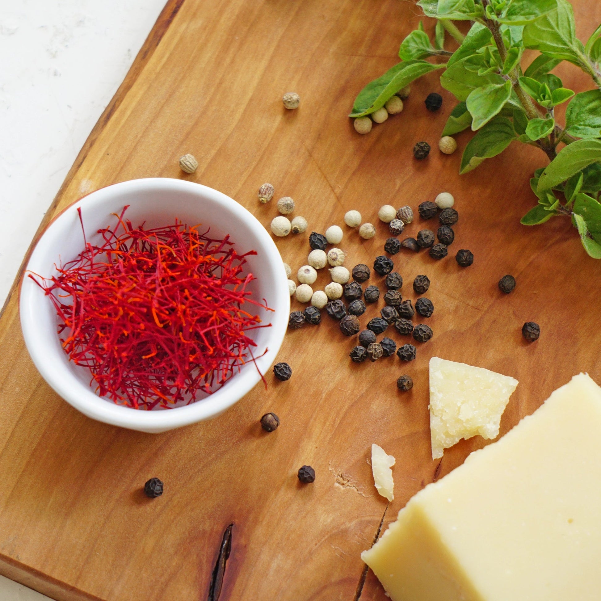 braunes Holzbrett mit Safranfäden in weisser Schüssel weisse und schwarze Kampot Pfefferkörner lose ein Stück Parmesan zwei Zweige Basilikum